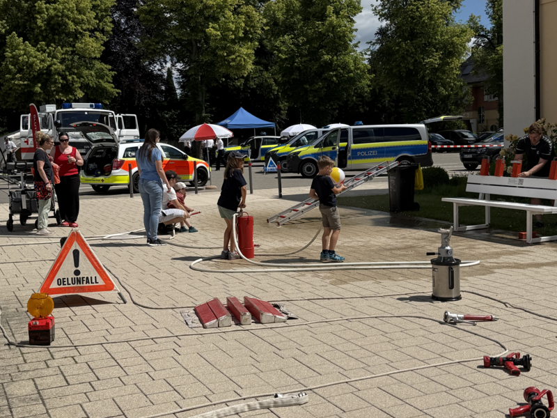 Vor einem Gebäude findet eine Übung mit Feuerwehr- und Polizeifahrzeugen statt, wobei Schläuche und ein Warnschild mit der Aufschrift "Ölunfall" auf dem Boden zu sehen sind.