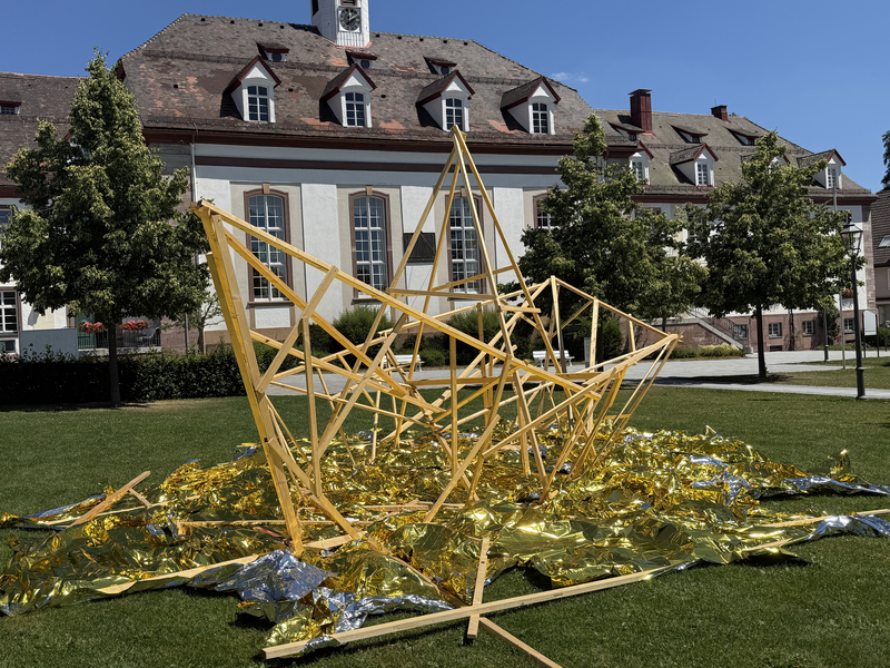 Vor einem historischen Gebäude steht eine skulpturale Installation aus dünnen Holzlatten und goldener sowie silberner Folie auf einem Rasen.