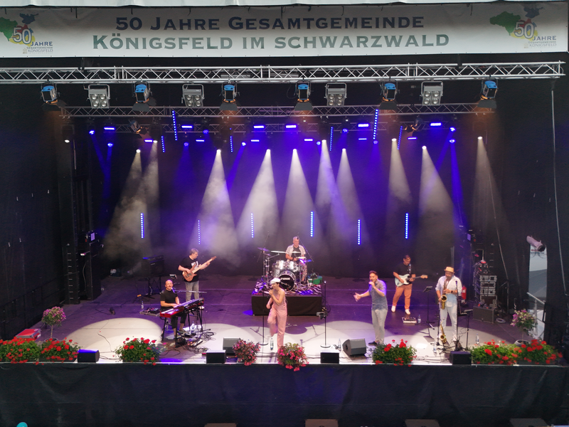 Eine Band tritt auf einer beleuchteten Bühne draußen auf, umgeben von Blumen, mit einem Banner, das ein Jubiläum der Gesamtgemeinde Königsfeld im Schwarzwald feiert.