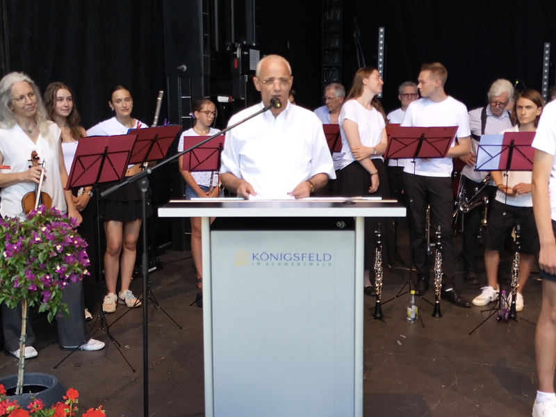 Eine Gruppe von Musikern steht auf einer Bühne, während ein Mann an einem Pult spricht, umgeben von blühenden Pflanzen und Notenständern.