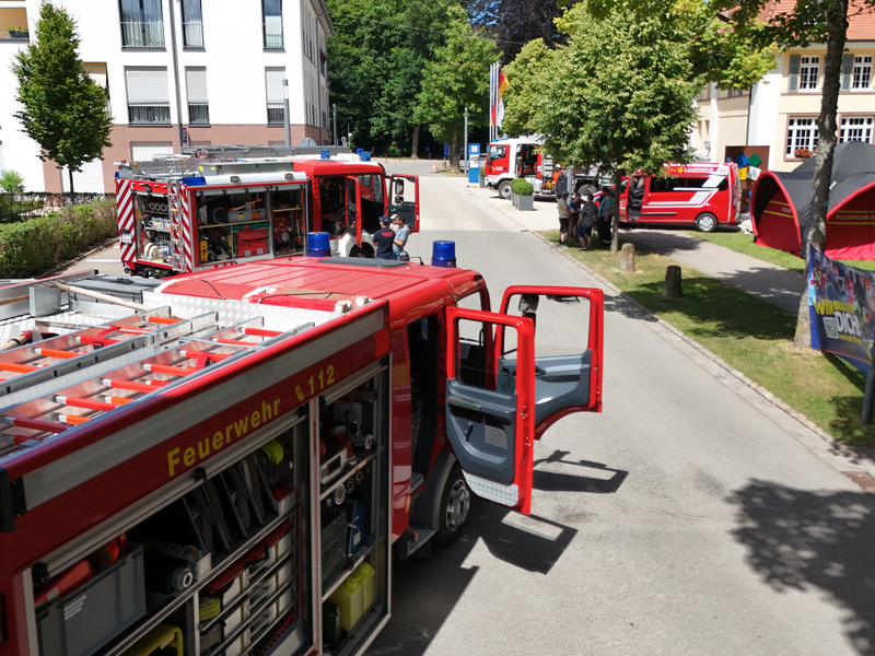 Auf einer Wohnstraße sind mehrere Feuerwehrfahrzeuge im Einsatz, umgeben von modernen Gebäuden und Bäumen.