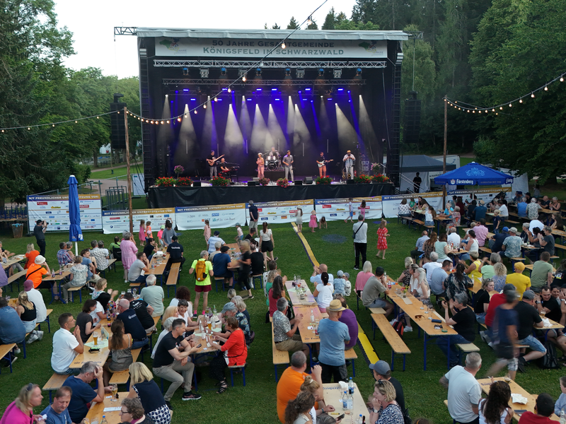 Eine große Menschenmenge sitzt an Biertischen im Freien und schaut auf eine Bühne, auf der eine Band unter buntem Licht spielt.