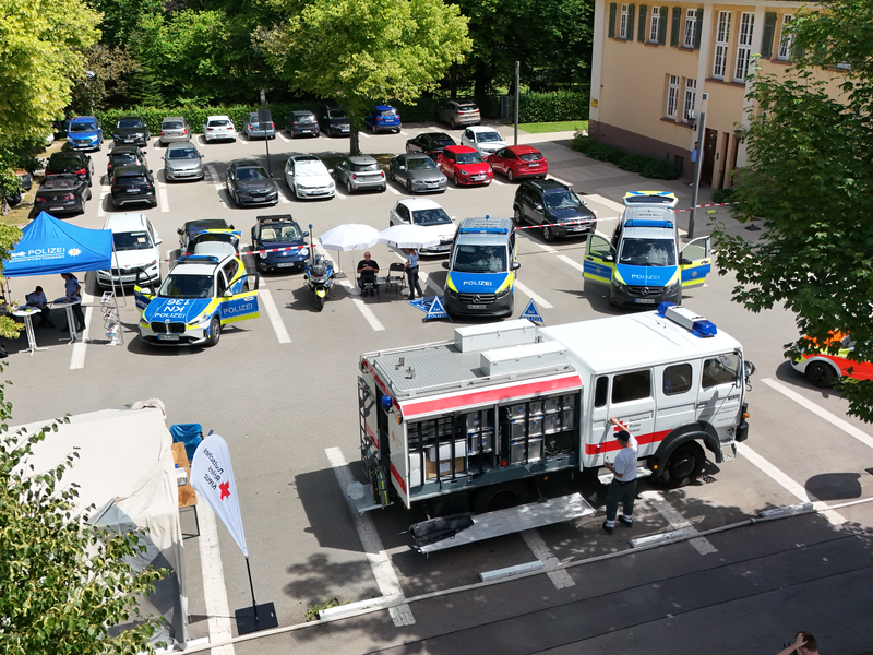 Auf einem Parkplatz stehen mehrere Polizei- und Rettungsfahrzeuge, umgeben von Pavillons und Menschen, die an einer Veranstaltung teilnehmen.