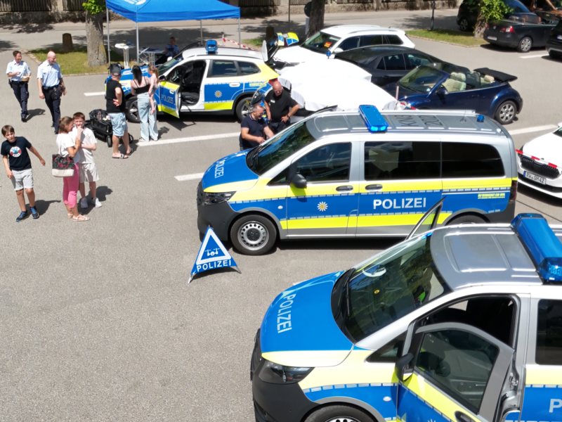 Auf einem Parkplatz stehen mehrere Polizei-Fahrzeuge umgeben von Menschen, die offenbar an einer Veranstaltung oder einem Treffen teilnehmen.