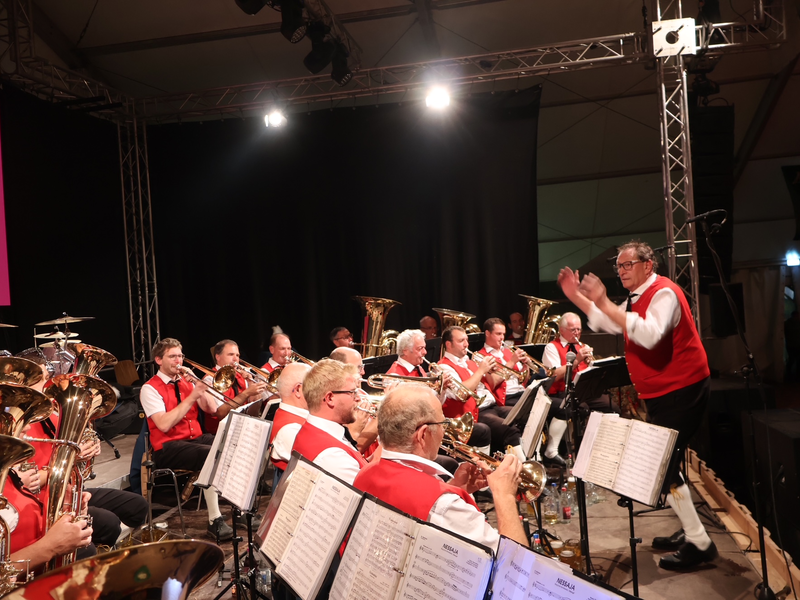 Eine Blaskapelle in roten Westen spielt auf einer Bühne, während ein Dirigent sie anleitet.