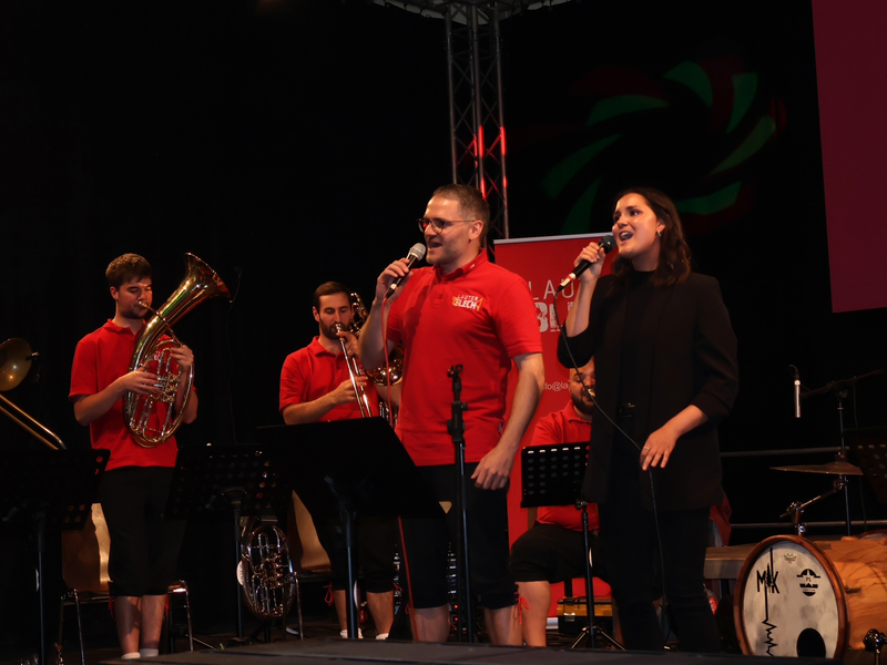 Eine Gruppe von Musikern, darunter eine Frau und ein Mann, singt auf einer Bühne, während andere Mitglieder Blechblasinstrumente spielen.