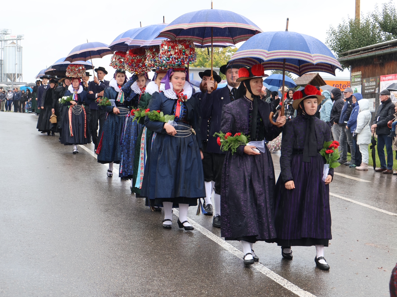 Eine Gruppe von Menschen in traditioneller Tracht, mit Hüten und Regenschirmen, marschiert auf einer regennassen Straße, während Zuschauer am Straßenrand stehen.
