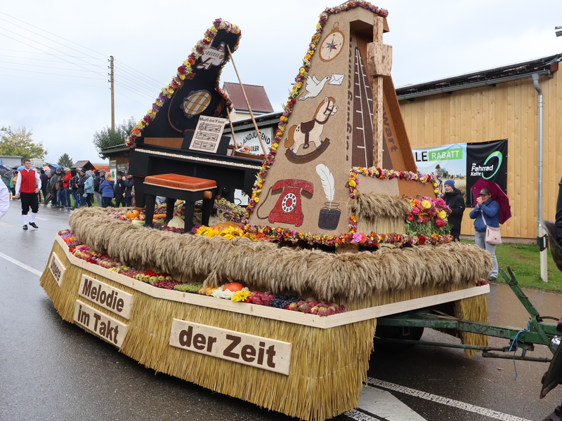 Ein bunter Festwagen, dekoriert mit Stroh und Blumen, zeigt ein Klavier und ein Pendel mit der Aufschrift "Melodie im Takt der Zeit" und wird von Menschen auf einer Straße begleitet.
