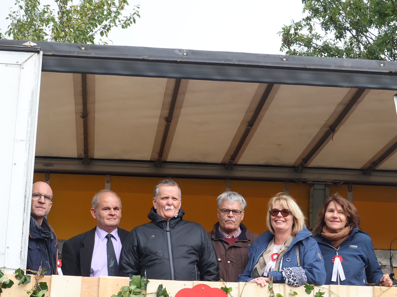 Sechs Personen stehen nebeneinander auf einer überdachten Bühne, die mit Pflanzen dekoriert ist, und lächeln in die Kamera.
