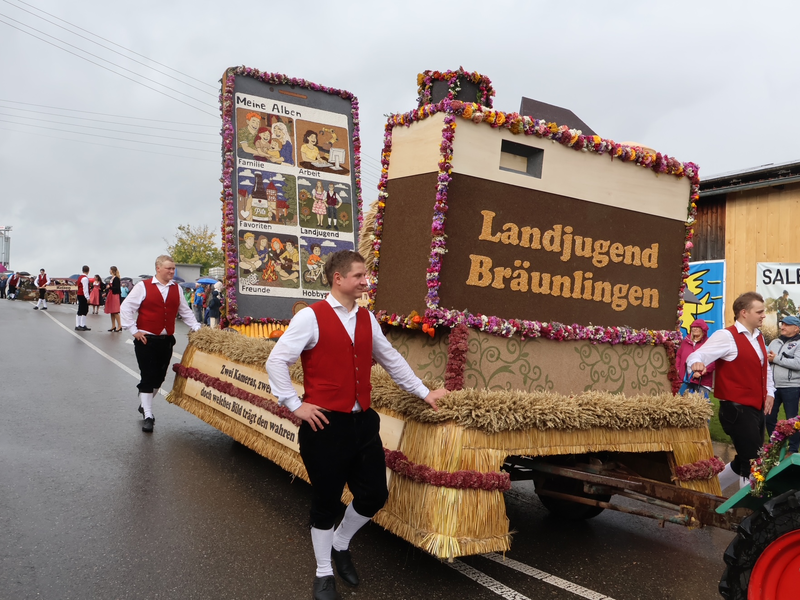 Ein festlich geschmückter Festwagen mit der Aufschrift „Landjugend Bräunlingen“ wird von Personen in traditioneller Kleidung über eine nasse Straße gezogen, umgeben von Zuschauern.