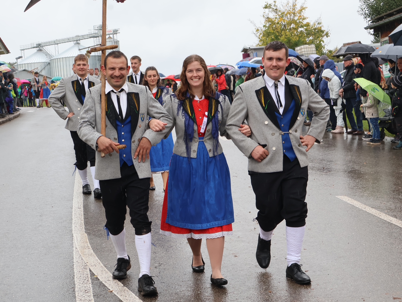 Eine fröhliche Gruppe von Menschen in traditioneller bayerischer Tracht läuft lächelnd bei einer Parade auf einer nassen Straße, während viele Zuschauer mit Regenschirmen zuschauen.