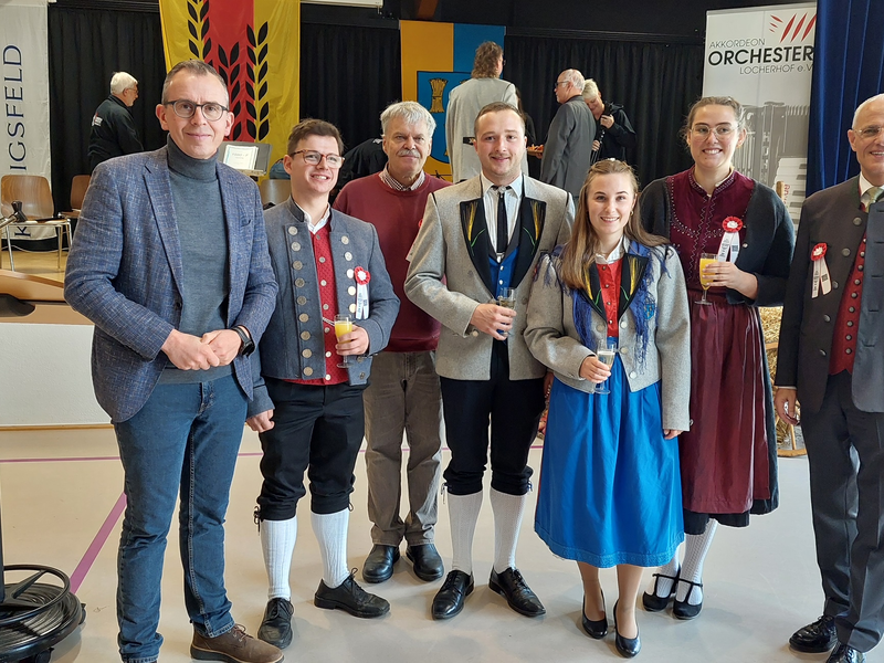 Eine Gruppe von sieben Personen, einige in traditioneller Tracht, steht in einer Halle vor Bannern und einem Rednerpult.