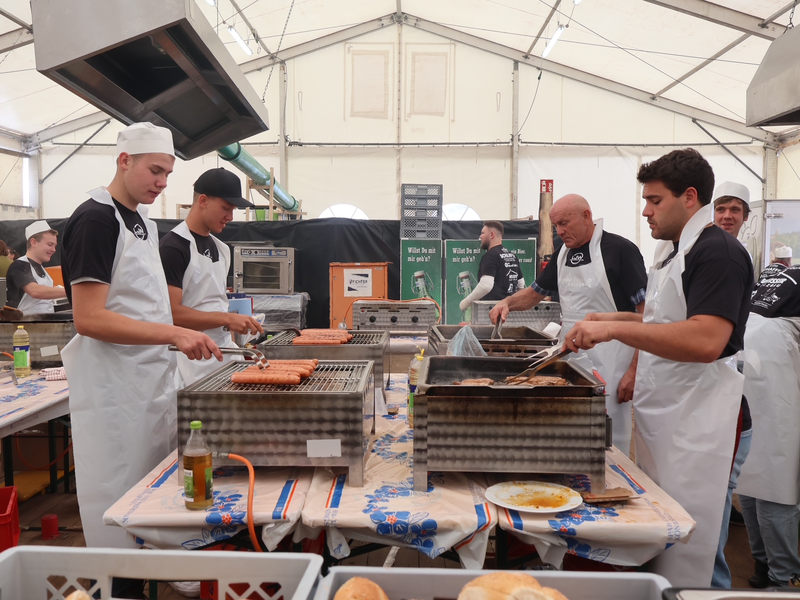 Mehrere Personen in Kochkleidung grillen in einem großen, hellen Zelt Würste und Fleisch.