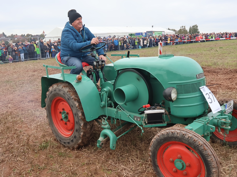 Ein älterer Mann fährt einen grünen Oldtimer-Traktor über ein Feld, während eine Menschenmenge im Hintergrund zusieht.