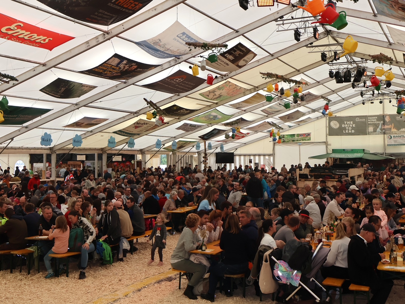 In einem großen Festzelt sitzen zahlreiche Menschen an langen Bänken und Tischen, dekoriert mit bunten Girlanden und Beleuchtung, während sie sich unterhalten und Getränke genießen.