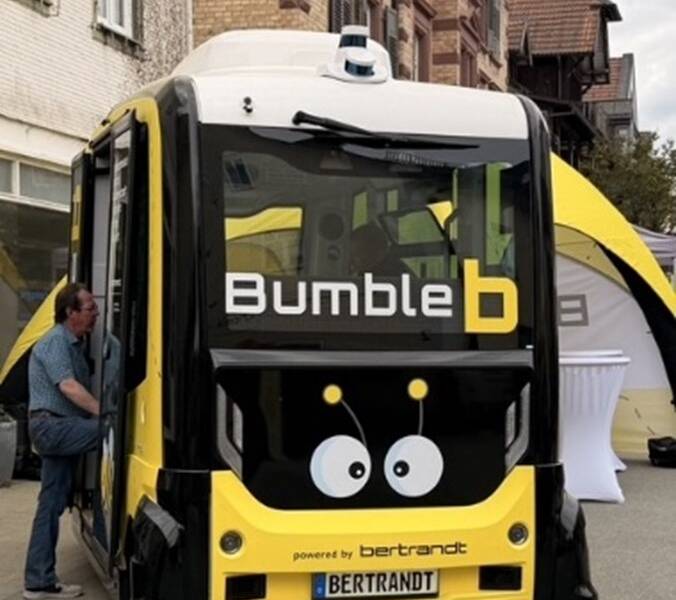 Ein selbstfahrender gelb-schwarzer Shuttlebus mit der Aufschrift "Bumble b" und einem Gesichtsmotiv auf der Vorderseite steht vor einem Gebäude, während eine Person an der Einstiegstür sichtbar ist.