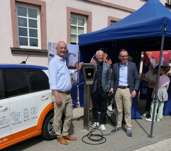 Drei Männer stehen vor einem geparkten Auto und einer Ladestation, neben einem blauen Pavillon.