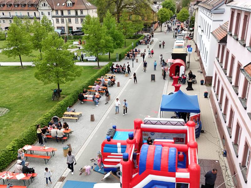 Eine städtische Straße ist für ein Straßenfest gesperrt, mit bunten Hüpfburgen, Zelten und Menschen, die an verschiedenen Ständen und Aktivitäten teilnehmen.