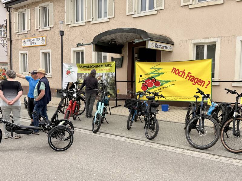 Vor einem Gebäude mit gelben Bannern sind mehrere Fahrräder und eine kleine Gruppe von Menschen versammelt.