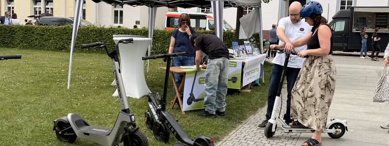 Zwei Personen mit Helmen stehen neben E-Scootern vor einem Zelt, während eine andere Person sich an einem Tisch unterhält.