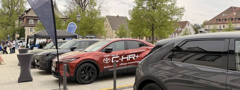 Mehrere parkende Autos stehen nebeneinander auf einem Parkplatz, umgeben von Bäumen und Gebäuden, mit Fahnen im Vordergrund.