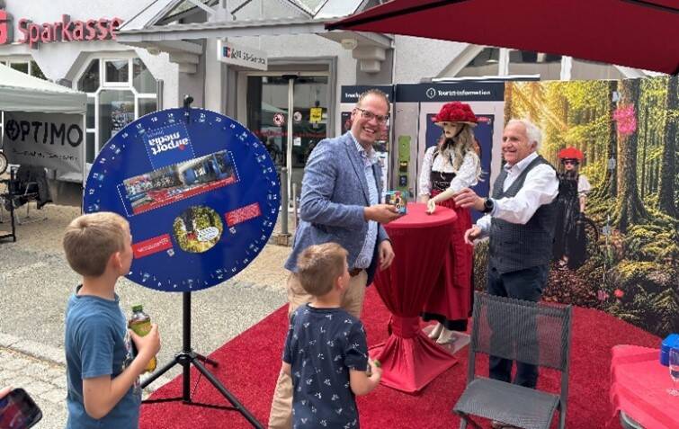 Ein Mann und ein Junge stehen vor einem Glücksrad auf einem roten Teppich, während ein anderer Mann neben einem Tisch mit einer bemalten Puppe steht.