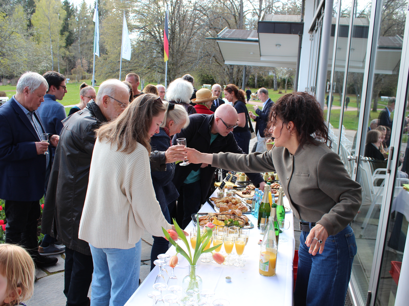 Eine Gruppe von Menschen versammelt sich bei einem Buffet im Freien, während eine Frau Getränke einschenkt.
