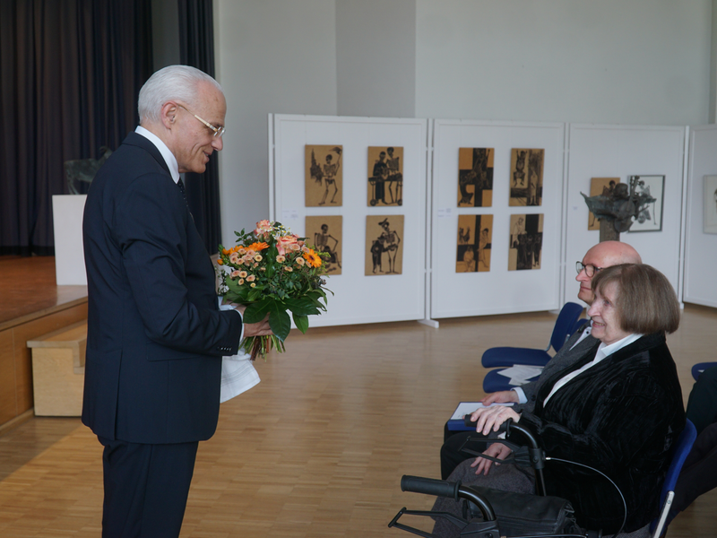 Ein Mann im Anzug überreicht einer sitzenden Frau einen Blumenstrauß in einem Ausstellungsraum mit Kunstwerken an der Wand.