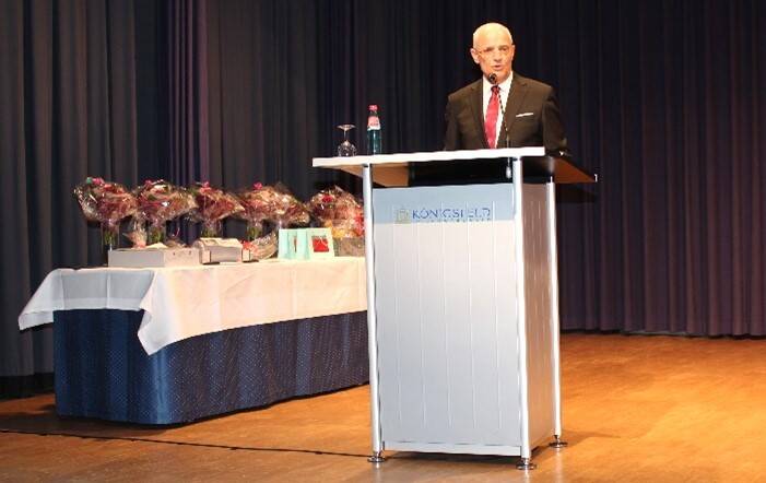 Ein älterer Mann steht an einem Rednerpult auf einer Bühne, während im Hintergrund ein Tisch mit mehreren Geschenken und Blumen zu sehen ist.