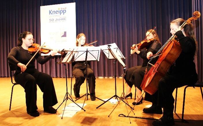 Vier Musikerinnen in schwarzer Kleidung spielen Streichinstrumente auf einer Bühne vor einem Vorhang.