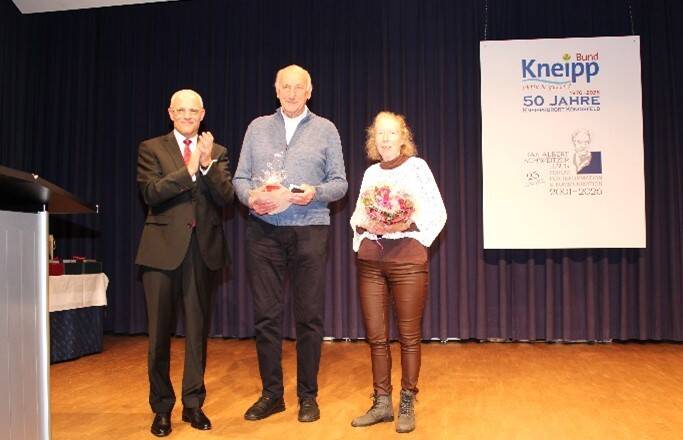 Drei Personen stehen auf einer Bühne; der Mann links trägt einen Anzug und klatscht, die beiden Personen in der Mitte und rechts halten jeweils Blumensträuße, und im Hintergrund ist ein Schild mit der Aufschrift "Kneipp" zu sehen.