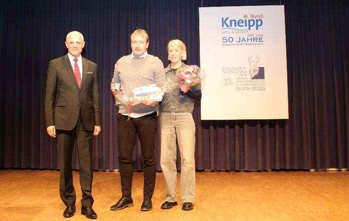 Drei Personen stehen auf einer Bühne vor einem blauen Vorhang, wobei der Mann links einen Anzug trägt und die zwei Personen rechts Blumensträuße halten.