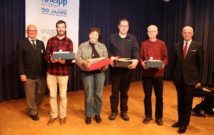 Sechs Personen stehen auf einer Bühne, von denen vier Geschenke in den Händen halten, während im Hintergrund ein Banner mit "50 Jahre Kneipp" zu sehen ist.