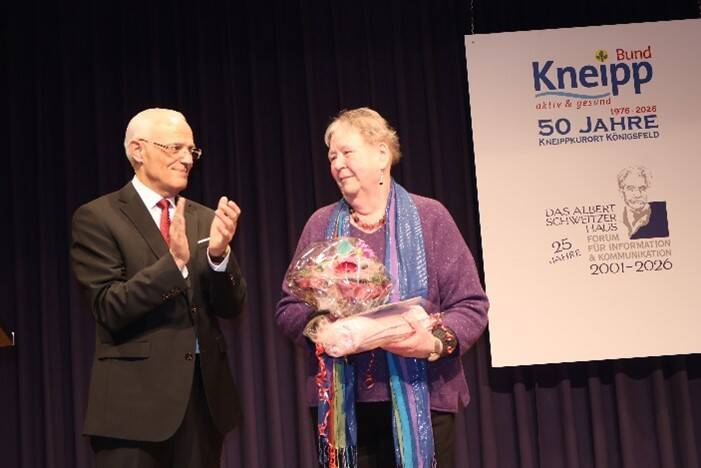 Ein älterer Mann in Anzug und Krawatte applaudiert einer älteren Frau, die einen Blumenstrauß hält, vor einem Plakat zur Feier von 50 Jahren Kneipp-Kurort Königsfeld.