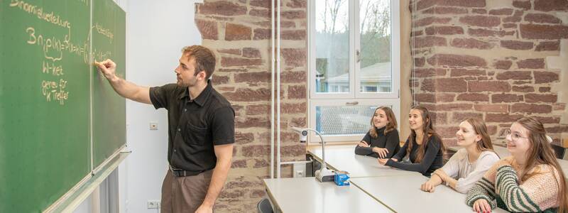 Ein Lehrer steht vor einer grünen Tafel und erklärt einer Gruppe von vier Schülerinnen eine mathematische Gleichung.