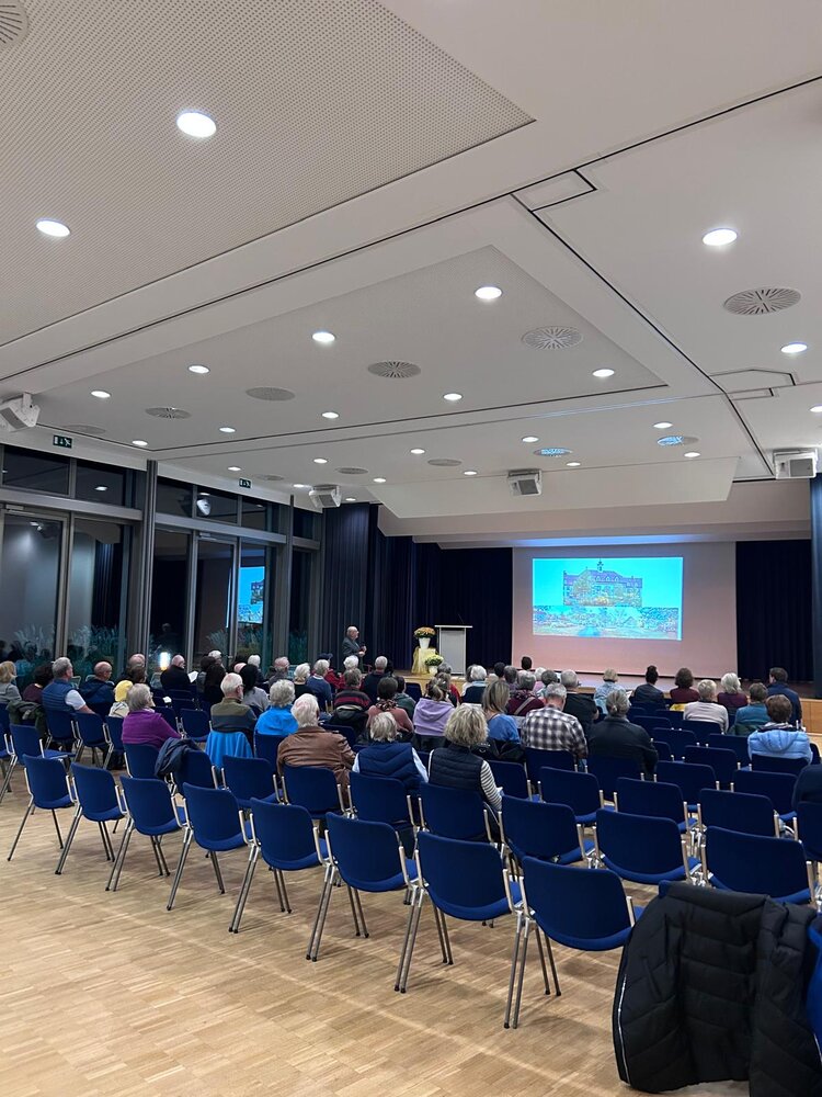 In einem Konferenzraum sitzen zahlreiche Menschen auf blauen Stühlen und schauen auf eine Bühne, auf der ein Vortragender neben einer Leinwand steht.
