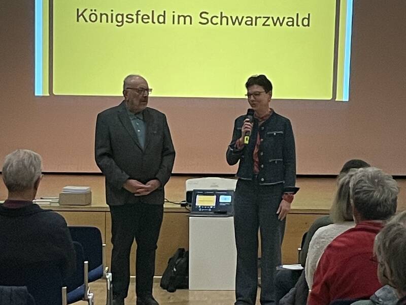 Zwei Personen stehen nebeneinander vor einer Projektionsleinwand mit dem Schriftzug "Königsfeld im Schwarzwald" und einem Publikum im Vordergrund.