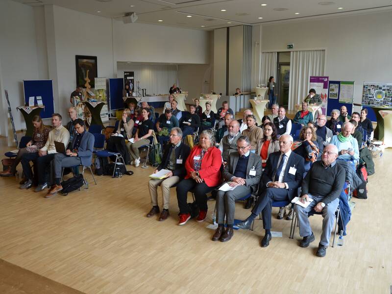 In einem Konferenzraum sitzen zahlreiche Menschen auf Stühlen und hören einem Vortrag zu.