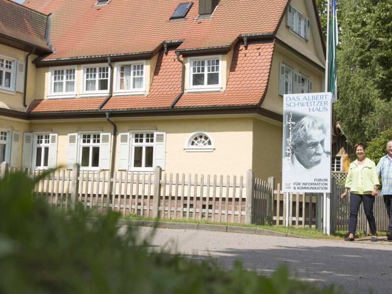 Eine Gruppe von fünf Personen geht an einem großen, zweistöckigen Gebäude mit rotem Dach und einem Schild mit der Aufschrift "Das Albert Schweitzer Haus" vorbei, das von einem Baum und Rasen umgeben ist.