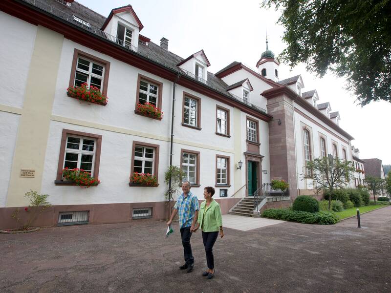 Ein Mann und eine Frau laufen händchenhaltend an einem älteren Gebäude mit Kirchturm vorbei.