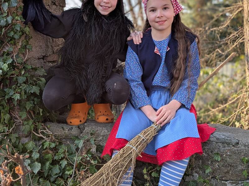 Zwei Kinder sitzen verkleidet auf einer mit Efeu bewachsenen Mauer, eines in einem schwarzen Kostüm mit Fliegerkappe und das andere in einem blauen Kleid mit Schürze, gestreiften Strümpfen und einer rot-weiß karierten Kopfbedeckung, während es einen Reisigbesen hält.