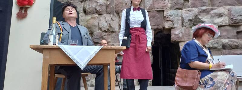 Drei Personen sind in einer Theaterszene zu sehen, wobei eine Person an einem Tisch sitzt und singt, eine als Kellnerin posiert und die dritte Person auf einem Hocker sitzt und Notizen macht, vor einem Hintergrund aus groben Steinen.