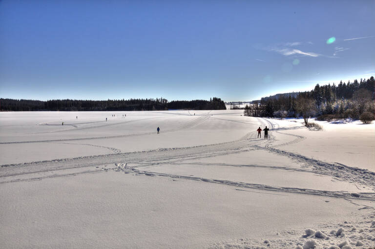 Verschneite Landschaft mit mehreren Menschen, die auf einem zugefrorenen See spazieren oder Ski fahren, umgeben von Wald unter klarem blauem Himmel.