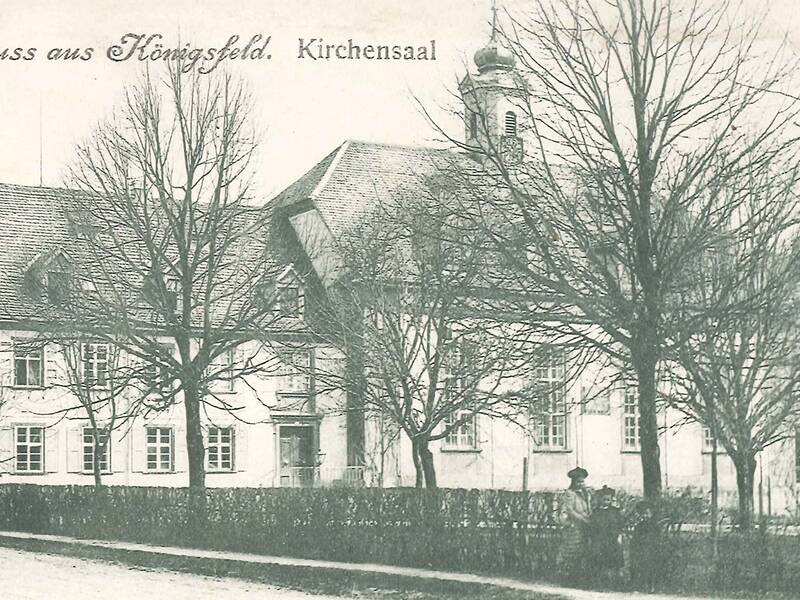 Schwarzweiß Bild mit kahlen Bäumen, im Hintergrund ein großes altes Gebäude, vorne zwei Spaziergämger ist historischer Kleidung