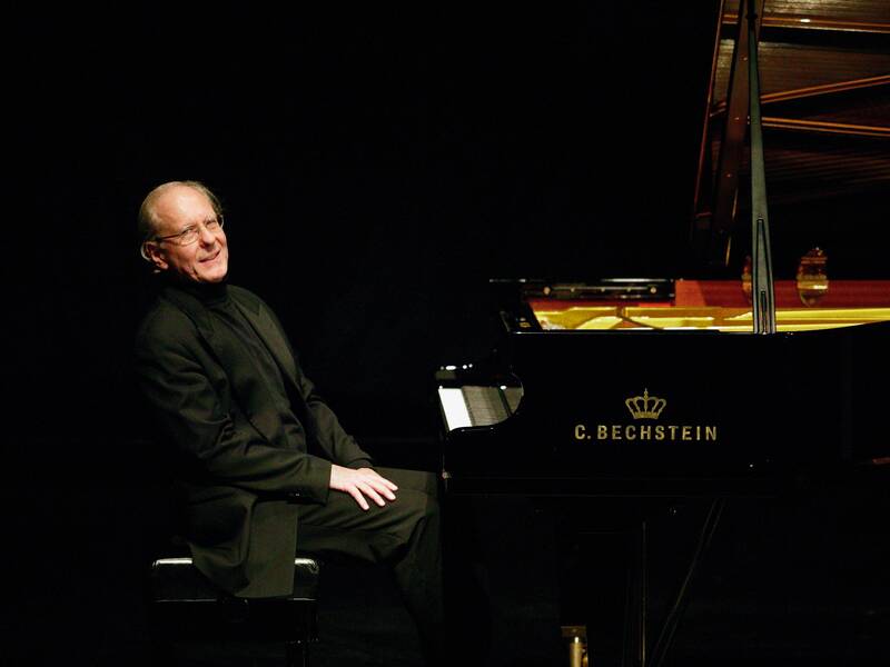 Ein älterer Mann sitzt lächelnd an einem schwarzen Flügel von C. Bechstein in einem gedimmten Raum.