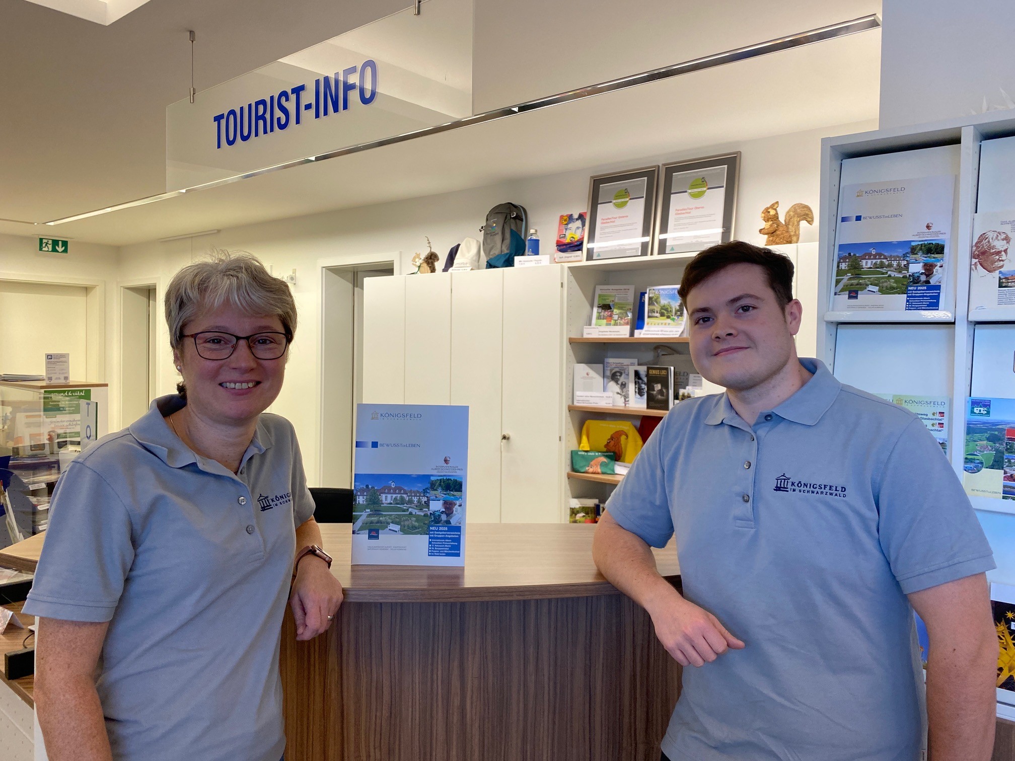 Zwei Personen in grauen Polohemden stehen am Tresen einer Tourist-Info, lächeln und wirken freundlich; im Hintergrund sind Broschüren und Auszeichnungen sichtbar.