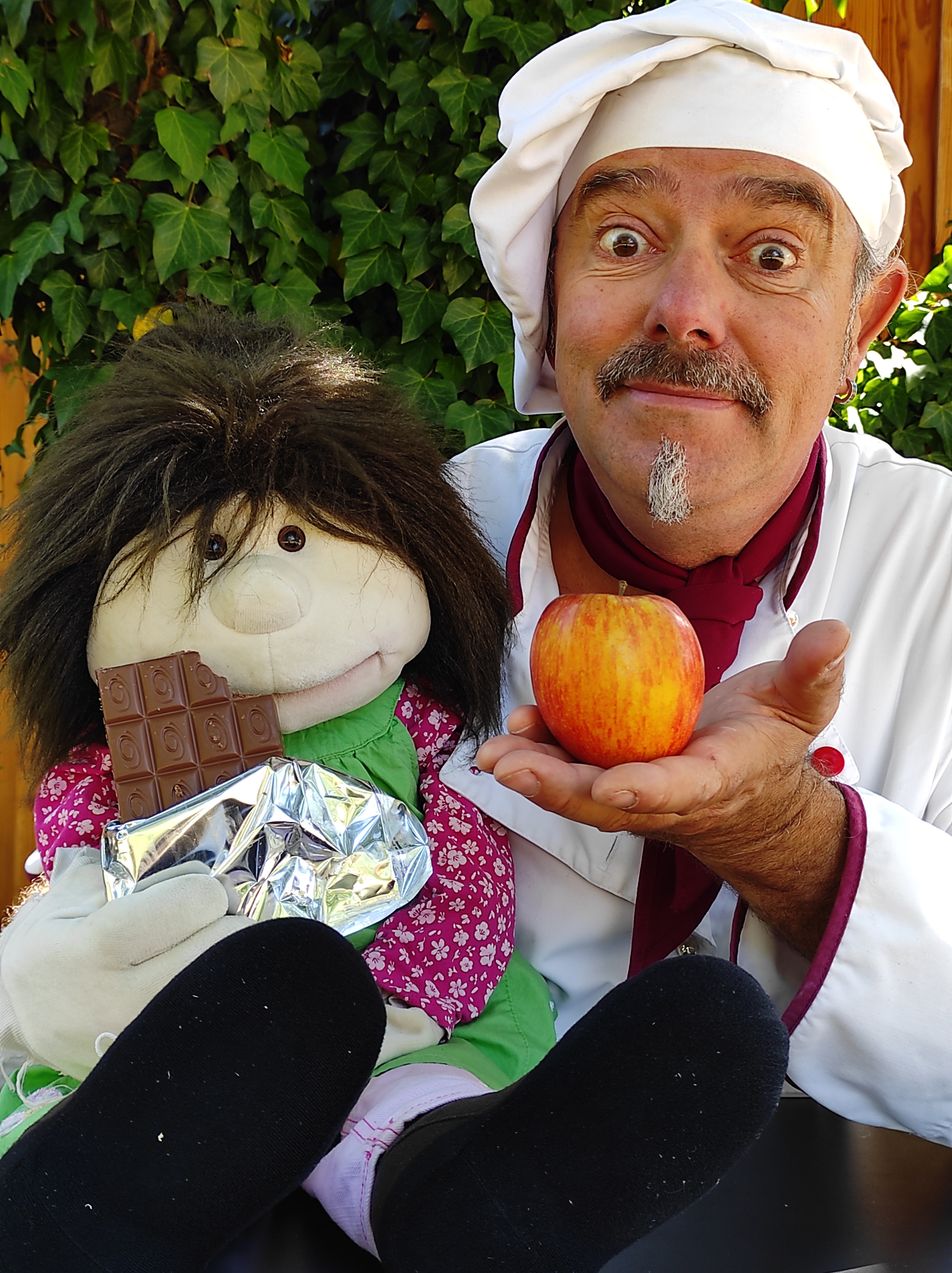 Ein Mann im Kochoutfit hält einen Apfel und sitzt neben einer Puppe, die eine halb ausgepackte Tafel Schokolade hält, vor einem Hintergrund aus grünen Blättern.