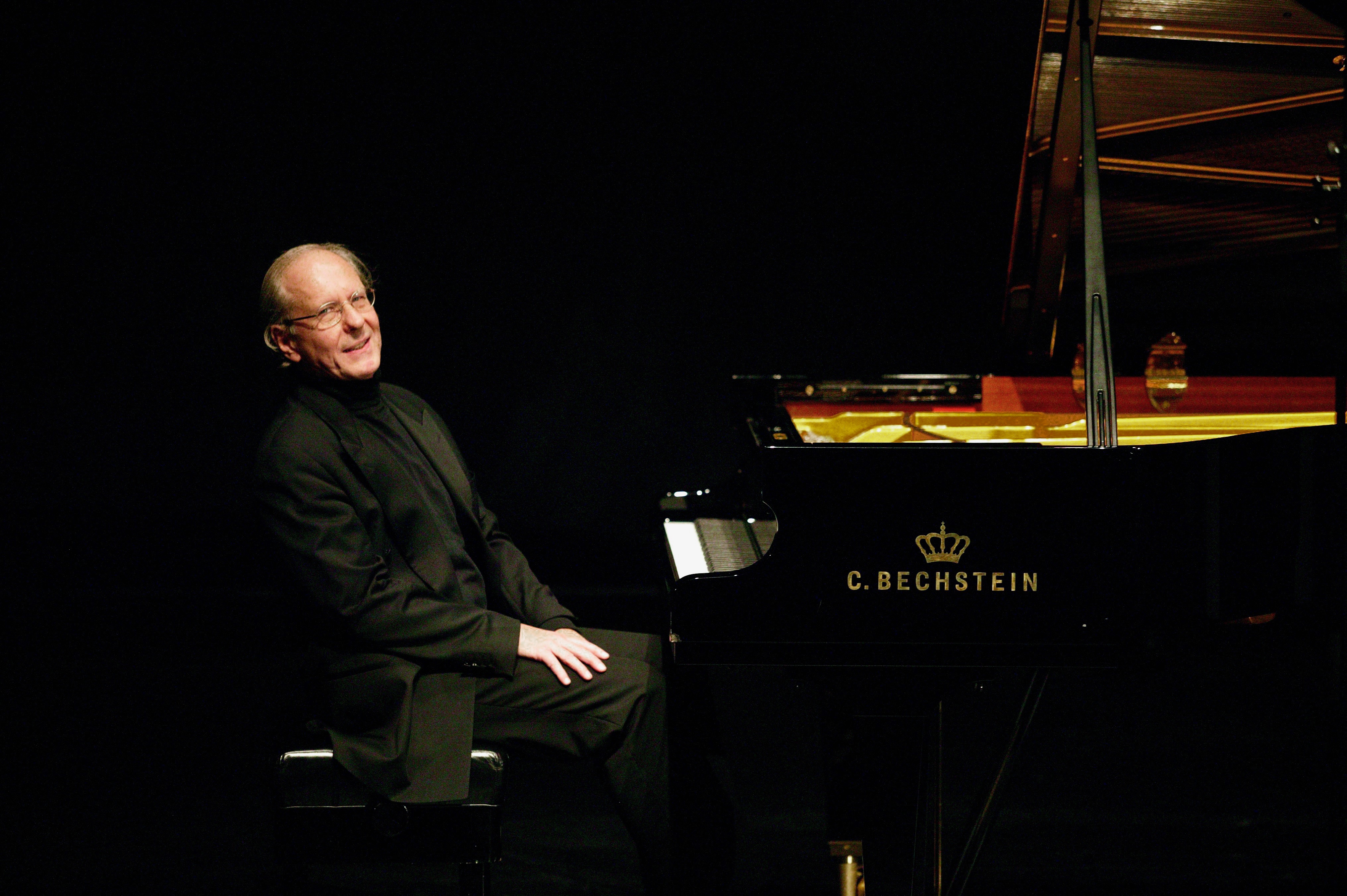 Ein älterer Mann sitzt lächelnd an einem schwarzen Flügel von C. Bechstein in einem gedimmten Raum.